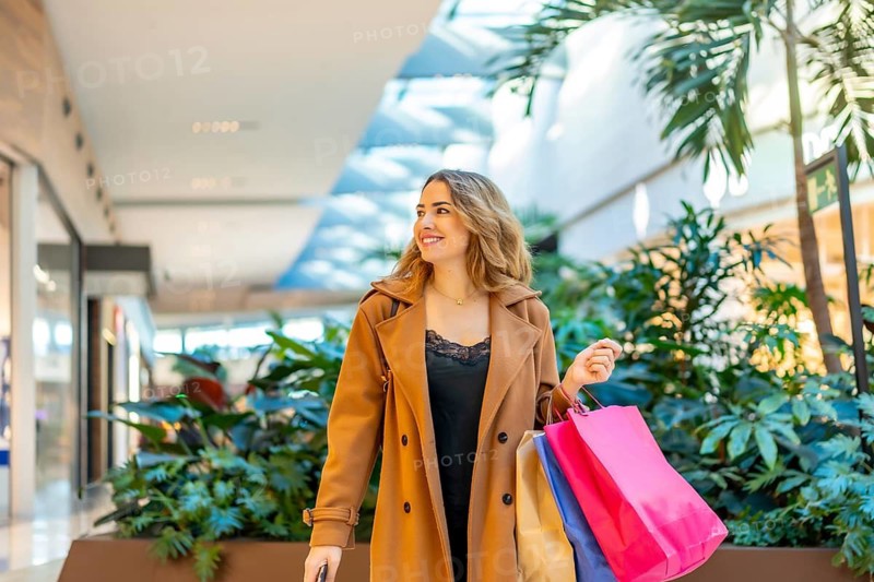 Young woman smiling while carrying colorful shopping bags. Walking through a bright. Contemporary shopping mall with lush green plants. Representing consumerism. Retail therapy. And a happy lifestyle