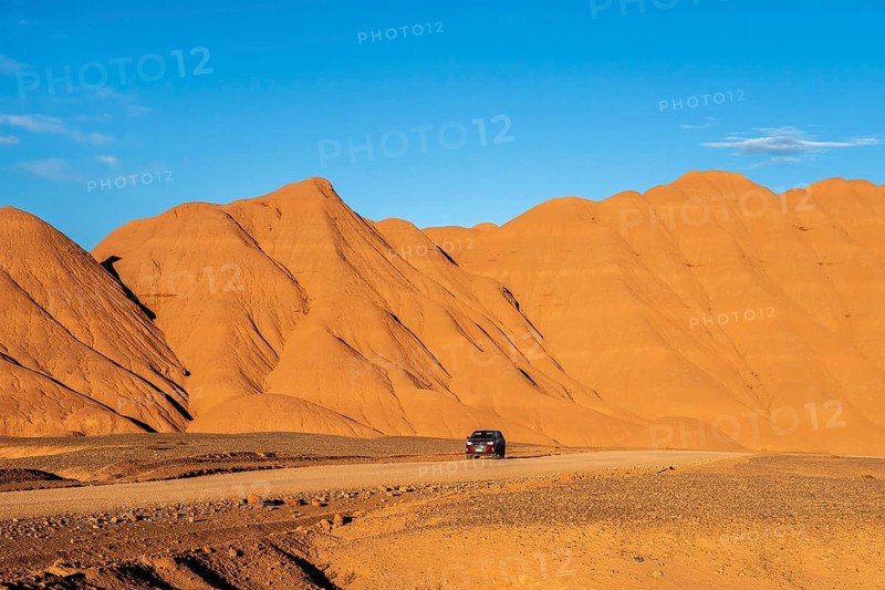 A vehicle traverses a winding road amidst the striking orange sandy mountains under a clear blue sky in La Puna, Argentina, showcasing the area's unique landscapes