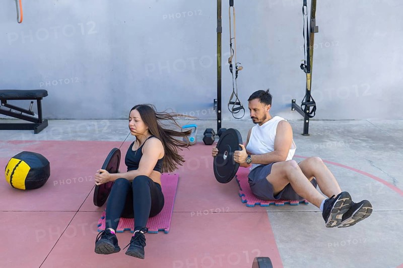 Two athletes are performing russian twists with weight plates, strengthening their core muscles in an outdoor gym setting, surrounded by various fitness equipment