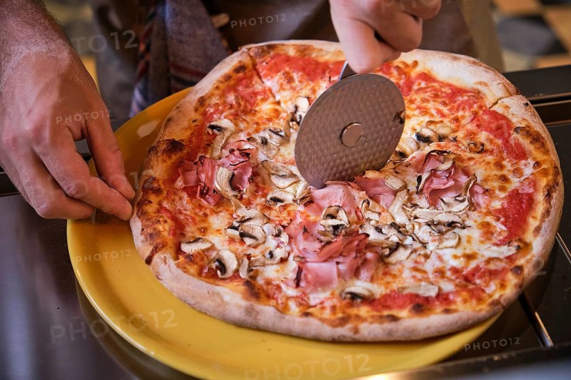 Cook cutting freshly baked pizza with ham and mushrooms using a round pizza cutter