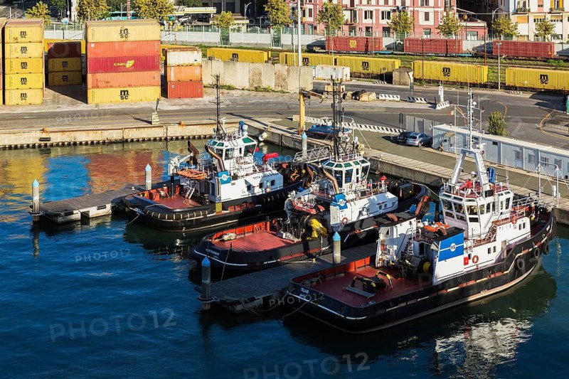 High angle view of docked red, white, blue tugboats, stacked yellow, reddish brown metal box shipping containers on dock, La Spezia cargo Port, Italy