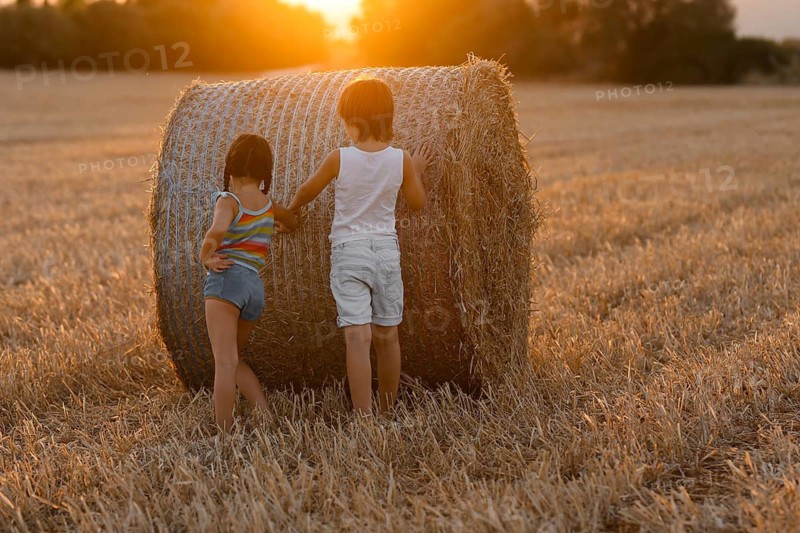 Back view of two unrecognizable children, a boy and a girl, play around a large hay bale in a field bathed in golden sunset light, capturing a moment of carefree, rural childhood