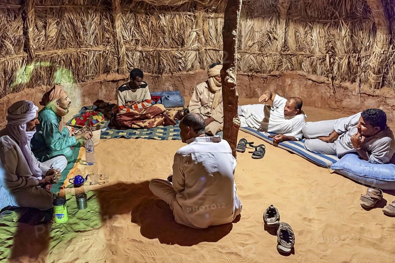 A group of people sit in a traditional hut with a sandy floor and relax in conversation, A group of Tuareg sit together in the evening and drink tea in the Sahara in Algeria