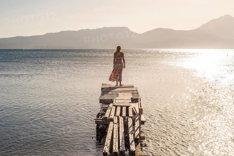 A woman stands on a rustic pier at Papikinou Beach, Milos Island, Greece, gazing at the tranquil sea and mountain silhouette during a stunning sunset