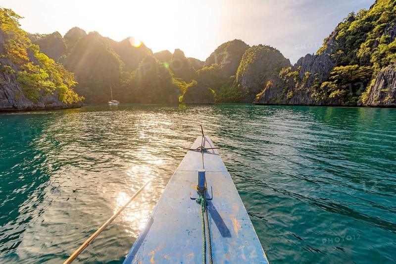 A view from a boat gliding over calm waters surrounded by lush, rocky mountains in the Philippines, bathed in the glow of the setting sun