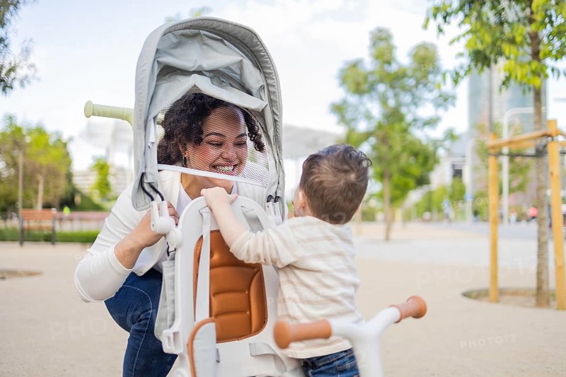 A joyful scene of a mother and her young child in a park, enjoying a sunny day. The mother smiles warmly as the child engages with her, surrounded by greenery