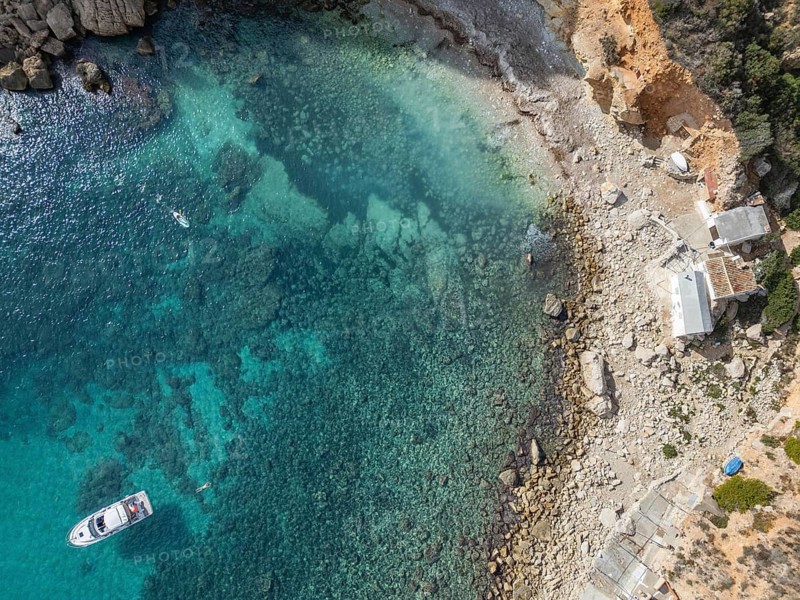 Aerial view of Costa Blanca's rocky coastline with a boat in the clear turquoise Mediterranean Sea. The coastal landscape captures the serene beauty of a sunny day by the sea