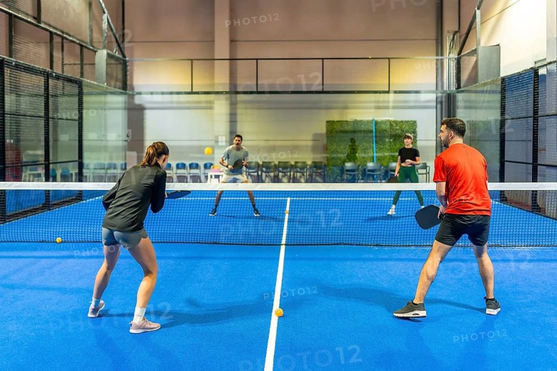 Four diverse players enjoying a competitive pickleball doubles match on a blue indoor court, actively swinging paddles at the ball while engaging in sport and fitness