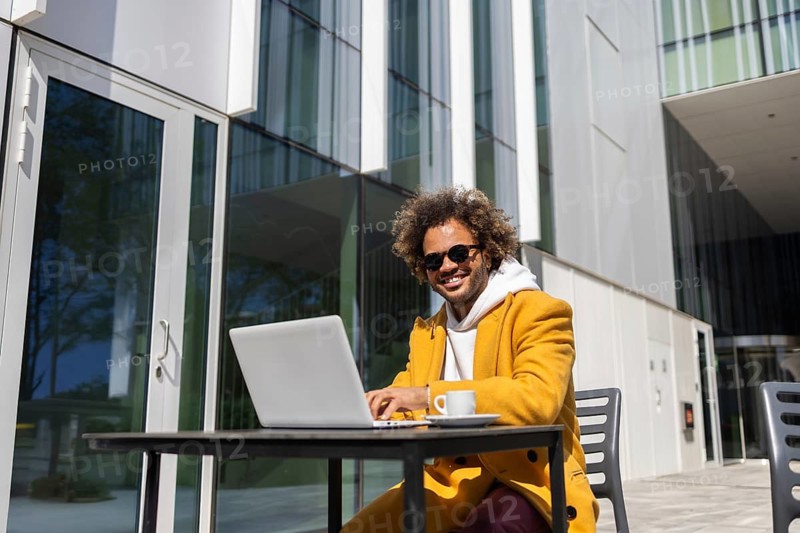 A businessman with afro hair and sunglasses sits happily at a table with a laptop and coffee outside a modern office building, showcasing a casual work environment