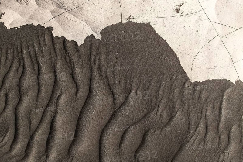 From above intricate textures of black sand shaped by natural forces on a beach. The ripples and patterns create a stark contrast against a lighter, cracked surface, showcasing nature's artistry