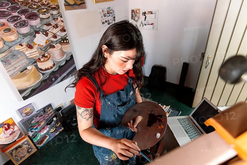 Young Latin woman focused on painting a canvas in her home art studio. Surrounded by paintings, she skillfully uses a palette, embodying creativity and artistic passion