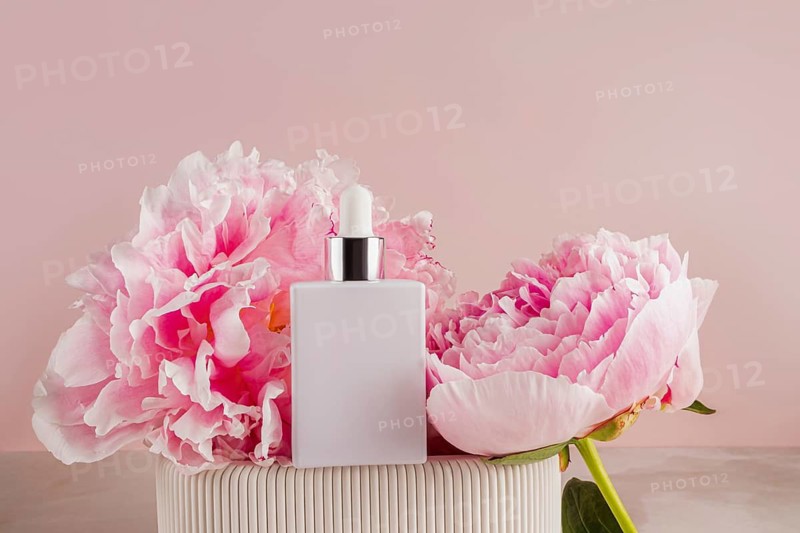 Elegant cosmetic bottle placed amidst two blooming pink peonies on a soft pink background. The scene evokes a sense of luxury and natural beauty, ideal for skincare products
