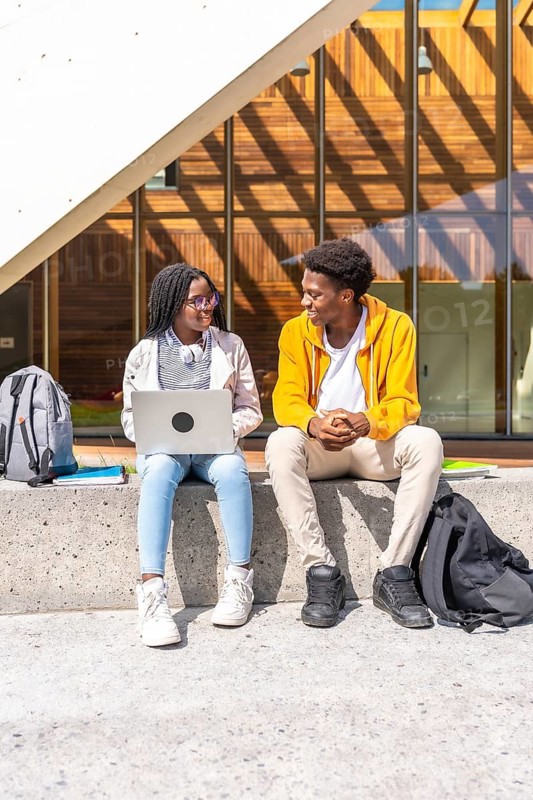 Vertical photo of two african students using laptop sitting outside the university building