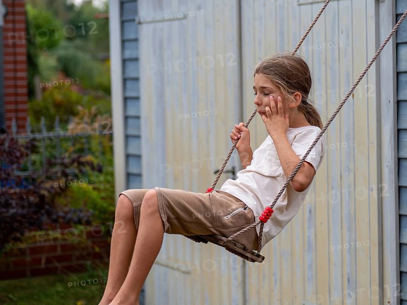 Boy swinging while sitting on children's swing at shed and garden gate, Germany