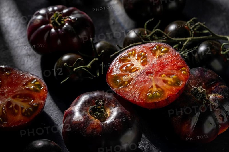 Black tomatoes, whole and sliced, display vibrant red interiors on a dark surface, illuminated by dramatic lighting. A taste of elegance and natural freshness