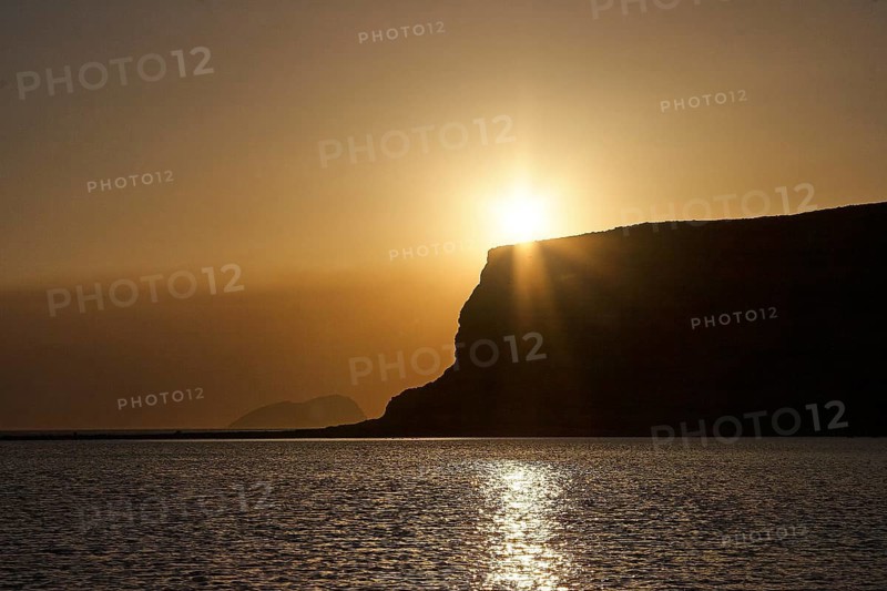 Dusk, sunset, sun as star, sun behind hill, bronze sky, cliff, island, sun reflection on water, Gramvoussa peninsula, Pirate's Bay, Balos, Tigani, West Crete, island of Crete, Greece