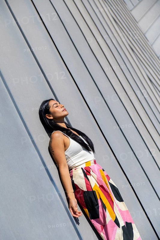 A woman in a vibrant, colorful outfit stands confidently against a backdrop of sleek modern architecture, highlighting the striking contrast between urban design and personal style