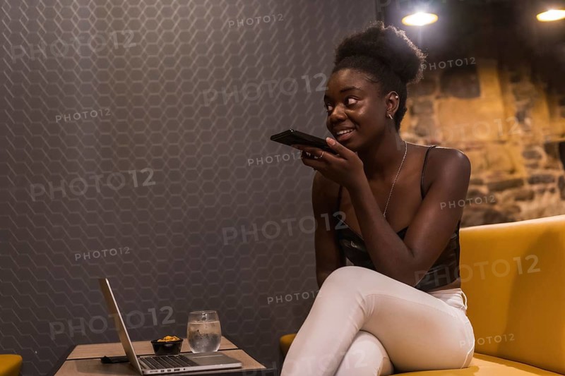 African woman sending a voice message sitting on a bar while working with laptop