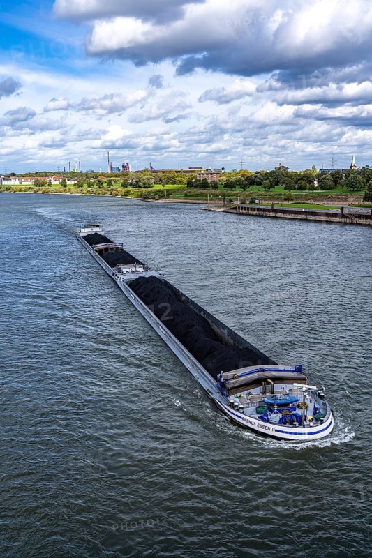 Rhine near Duisburg-Laar, freighter loaded with coal, houses on the dyke road, industrial backdrop of the ThyssenKrupp Steel steelworks in Bruckhausen, North Rhine-Westphalia, Germany