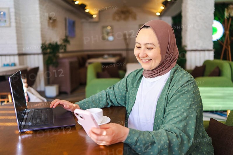A Muslim woman in a hijab is seated at a cafe table with a laptop, holding a phone, and smiling. The cozy cafe environment serves as her workspace
