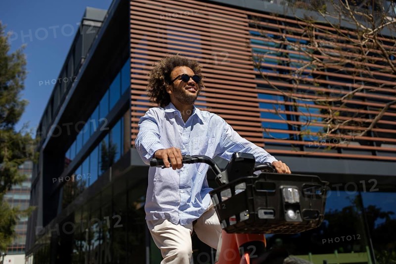 A relaxed man with sunglasses rides a public bicycle outside a modern building, enjoying a break from business. The sunny day complements his carefree and casual demeanor