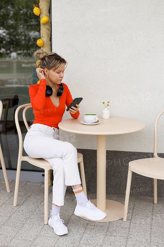 Young woman in bright orange top, sitting at an outdoor cafe in Warsaw She is engaged with her smartphone and headphones, alongside a cup of coffee on the table