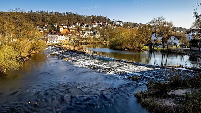 Small waterfall in a river in front of a village with autumnal trees and a clear sky, The weir of the Fulda in Melsungen