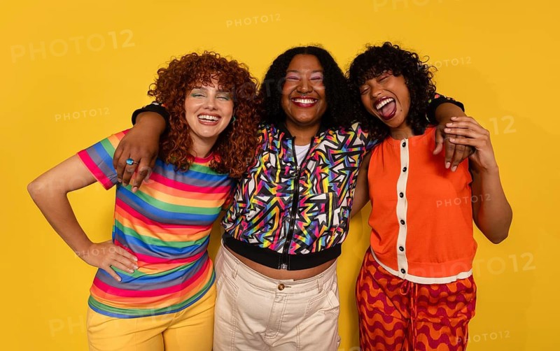 Three friends smile brightly in a colorful and cheerful setting. Standing against a vivid yellow background, they display camaraderie and joy with their vibrant outfits and joyful expressions