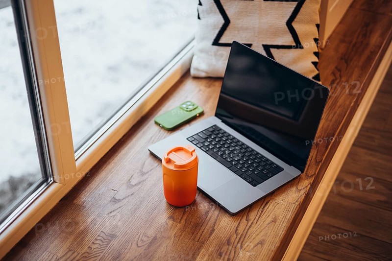 A cozy remote work setup featuring a laptop and a bright orange thermos cup beside a snowy window, reflecting a warm, productive workspace