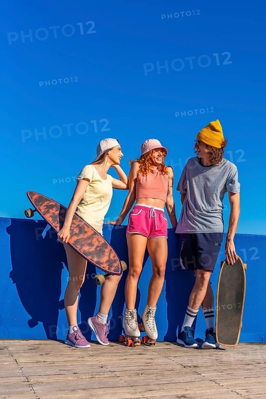 Three cheerful young friends with skateboards and roller skates are enjoying a sunny day, chatting and having fun against a vibrant blue wall