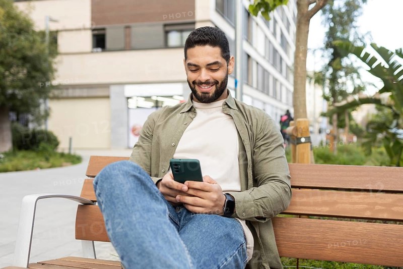A man sits on a park bench, smiling as he looks at his smartphone. Dressed casually, he enjoys a relaxed moment outdoors, surrounded by urban greenery