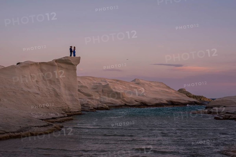 Unrecognizable couple standing on the unique white rock formations of Sarakiniko beach at sunset, with the tranquil sea and dusk sky in the background, on the island of Milos, Greece