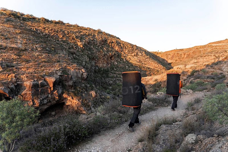 Two climbers carry crash pads along a rocky path surrounded by stunning natural scenery, preparing for a boulder climbing adventure in a remote, sunlit canyon