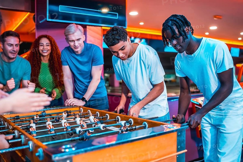 Multi ethnic friends engaging in an exciting foosball game at a lively entertainment center, filled with vibrant lights and laughter
