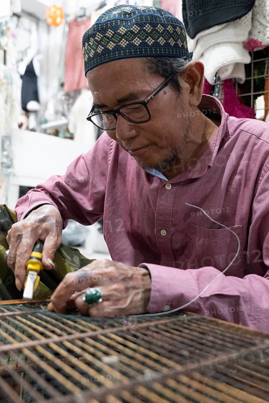 A balinese artisan wearing glasses works on a detailed wire project. His hands hold a tool, showcasing craftsmanship in a well lit workshop