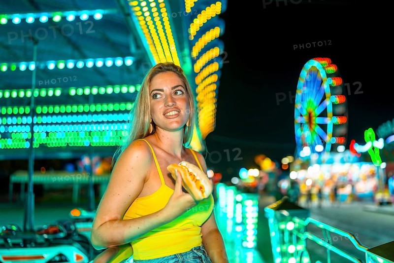 Blonde woman holding an unhealthy hot dog in a night fair in summer
