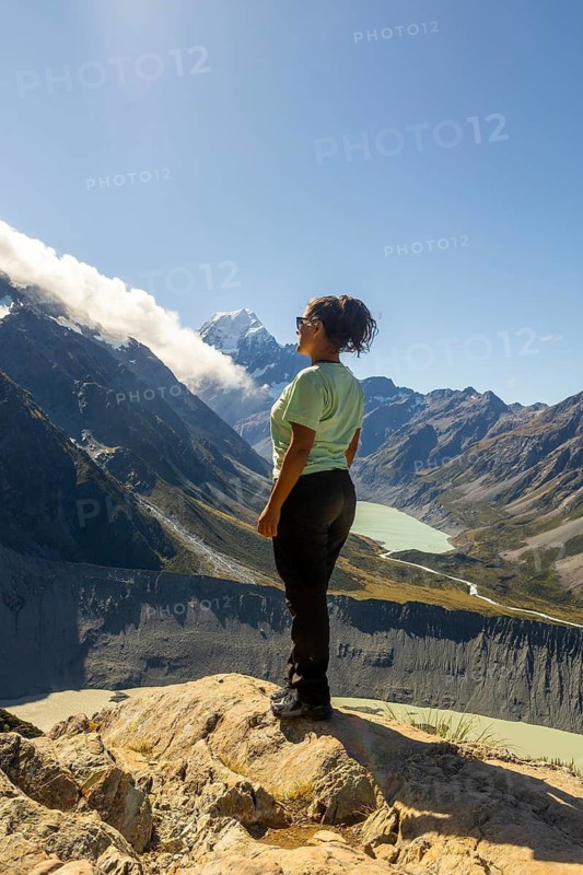 A woman stands on a rocky outcrop, taking in the breathtaking autumn views of Mount Cook in New Zealand. The clear blue sky enhances the majestic mountain scenery