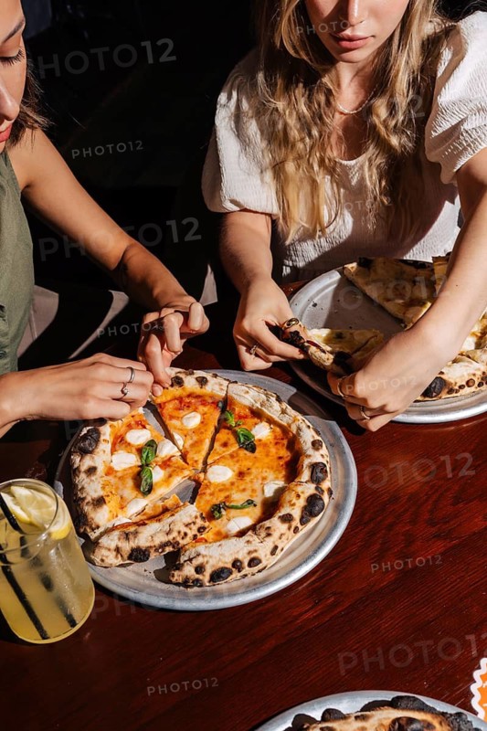 Two people share a Margherita pizza topped with mozzarella, basil, and tomato sauce. A refreshing lemonade sits on the table, enhancing the friendly dining experience