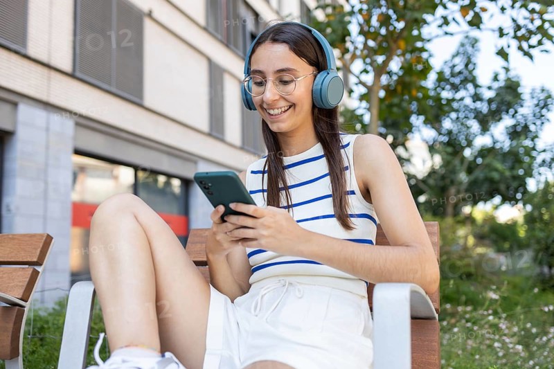 A woman sits outdoors in the city, listening to music on headphones while smiling at her phone. Her casual outfit and relaxed demeanor reflect a modern urban lifestyle
