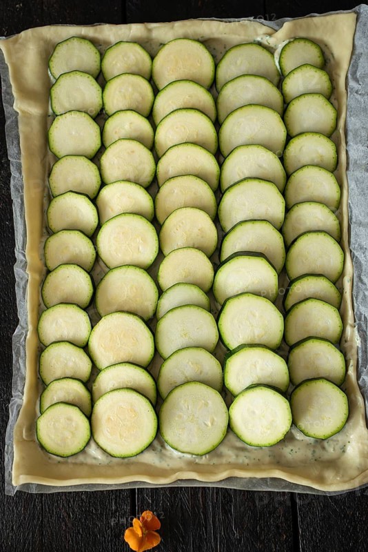 A visually appealing tart with neatly arranged zucchini slices on a pastry base, accompanied by a vibrant edible flower, highlighting fresh and natural ingredients