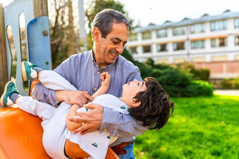 Grandfather holding his grandson playing on a playground slide, enjoying a sunny day in the park