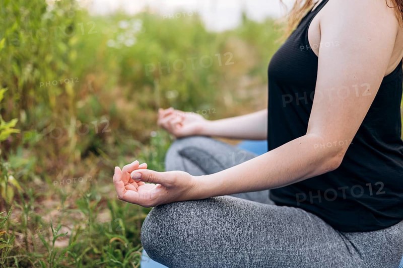 Cropped unrecognizable woman in a yoga pose meditating outdoors in a field, sitting cross-legged and focusing on mindfulness and relaxation amidst natural surroundings