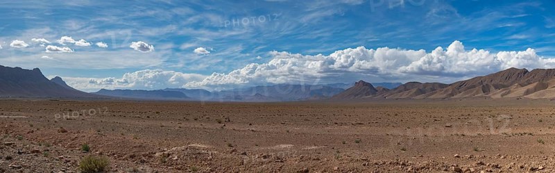 A vast desert landscape stretches out under a vibrant blue sky, with towering mountains on the horizon of High Atlas, Morocco. Sparse vegetation dots the foreground, adding depth to the scene
