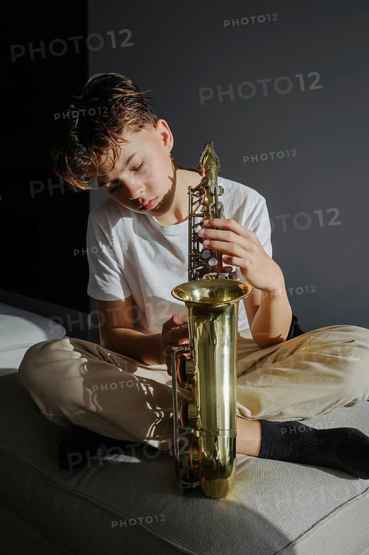 A young boy sits on a couch, holding a saxophone, bathed in soft natural light. His expression is thoughtful, reflecting a deep connection with music