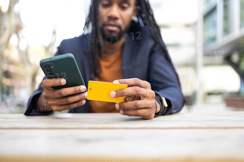 A cuban businessman sits at a wooden table outdoors, holding a smartphone and a yellow card, engaging in an online transaction or payment process under natural lighting