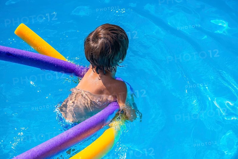 Young boy playing in a bright blue pool, smiling and floating on colorful pool noodles while splashing and enjoying sunny summer leisure and carefree vacation fun