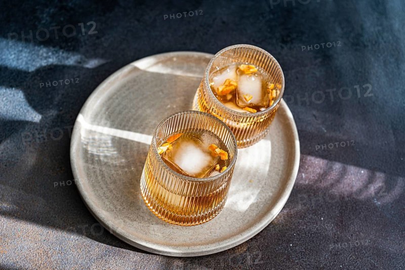 Top view of two spiced cognac based cocktails with cinnamon and star anise, served on a textured ceramic plate accompanied by almonds, peanuts, and walnuts