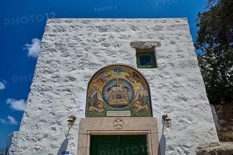 A white building with a green door and an ornate mosaic on a stone wall under a clear sky, evangelist symbols, lion, bull, eagle, lamb, Monastery tis Apokalipsis, Patmos, Dodecanese, Greek Islands, Greece