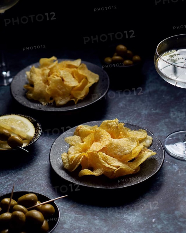 A close-up of golden potato chips on rustic black plates. Accompanied by green olives and sliced lemon, this elegant snack setup is perfect for a gourmet appetizer