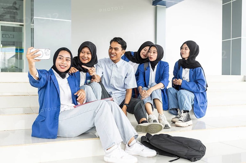 A diverse group of students, including Asian Muslim women, take a selfie on the steps of a school campus, sharing smiles and creating memories on their first day back to school